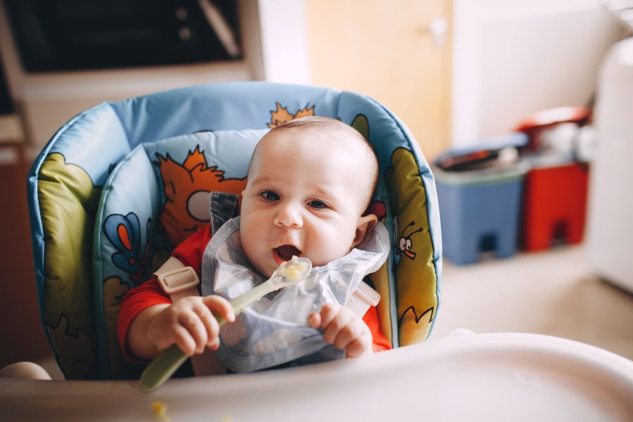mother feeding her baby first solid foods with a spoon in a high chair